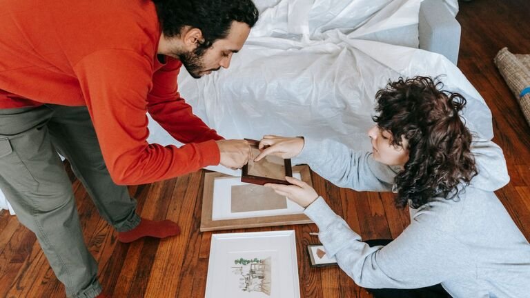 Pareja preparando cuadros enmarcados en el suelo junto a un sofá cubierto con una funda protectora antes de la mudanza